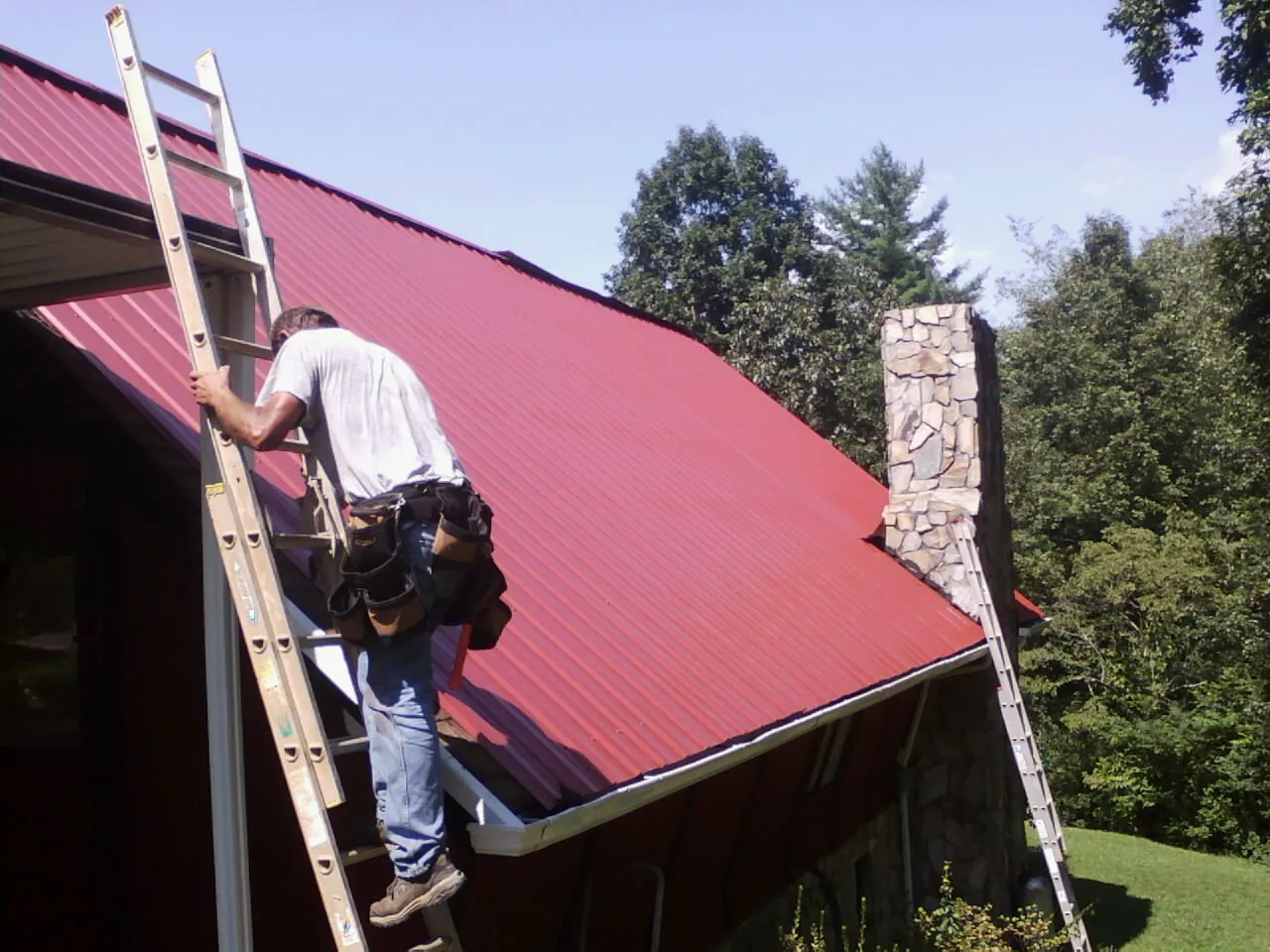 Roofer on a ladder beside a red metal roof and stone chimney — Soffit & Fascia Repair by Roofing Brothers in Levels